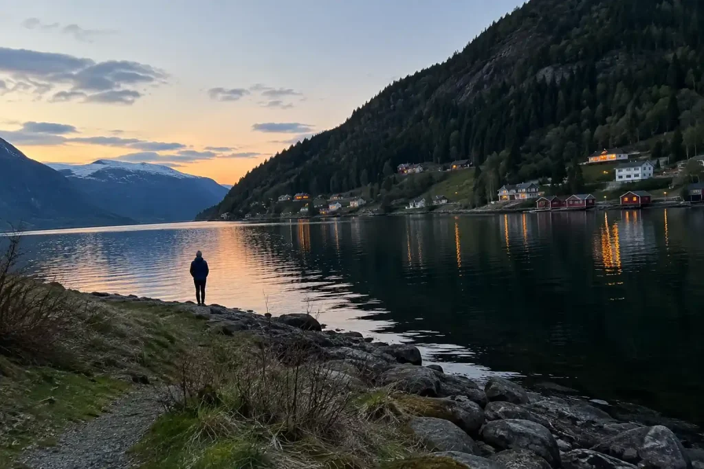 Skymning vid en tyst fjord med trähus och en ensam person vid strandkanten, i stämning från Döden vid fjorden på TV4.